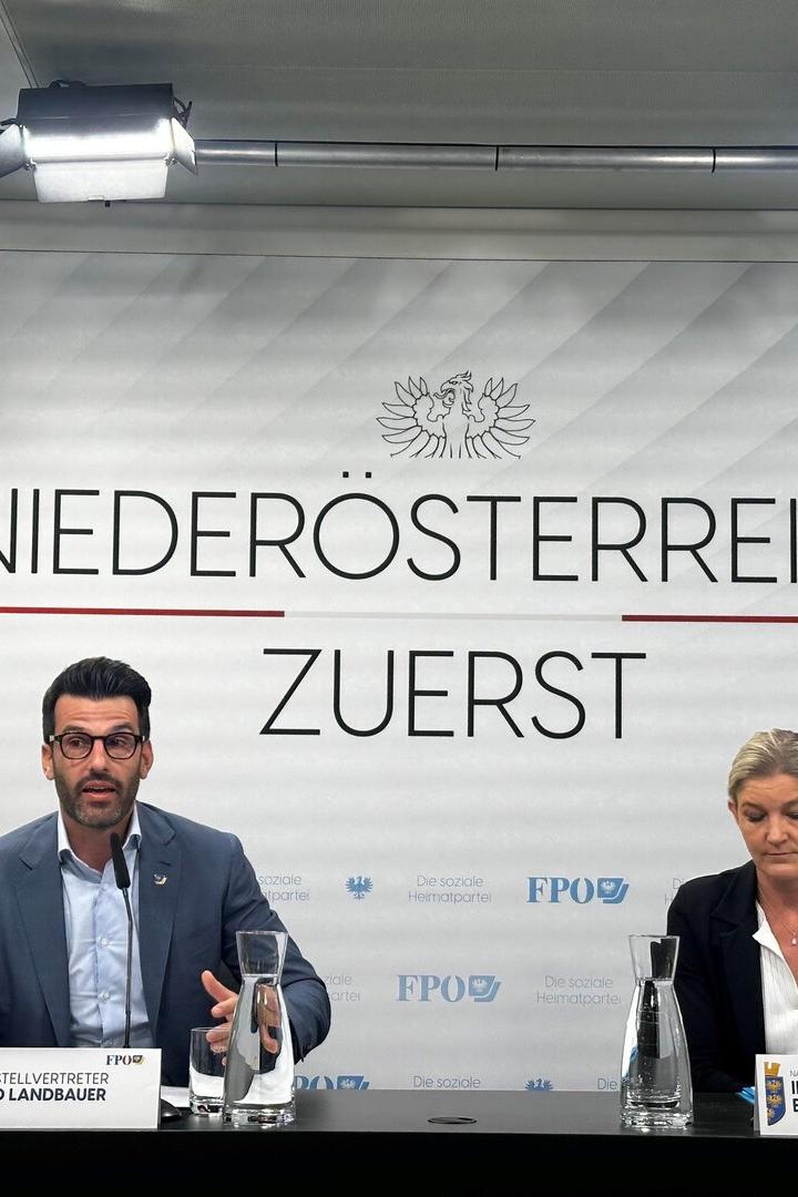 Zwei Personen sitzen an einem Tisch mit Namensschildern vor einer Wand mit der Aufschrift „Niederösterreich zuerst“.