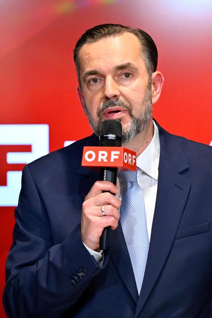 Mann im Anzug spricht in ein ORF-Mikrofon vor rotem Hintergrund.