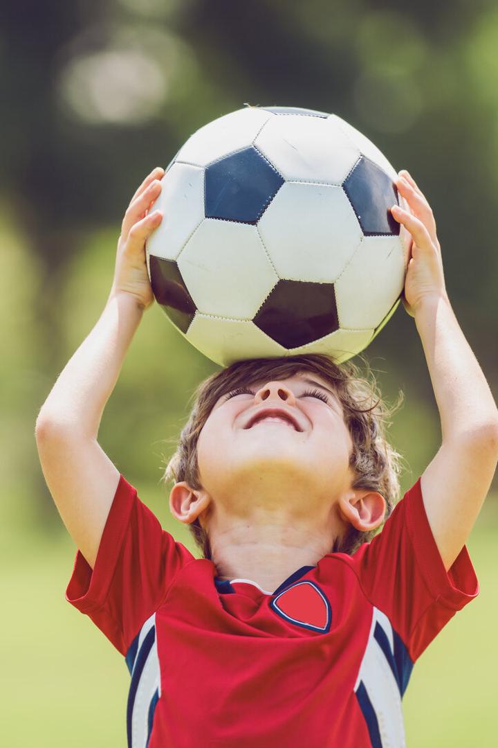 Ein Junge im roten Trikot hält einen Fußball über dem Kopf im Freien.