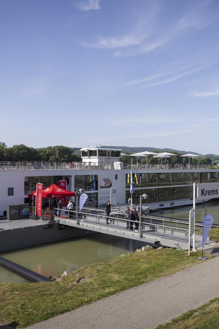 Anlegestelle am Fluss mit zwei Kreuzfahrtschiffen, Steg und Passagieren vor grünen Hügeln unter blauem Himmel.