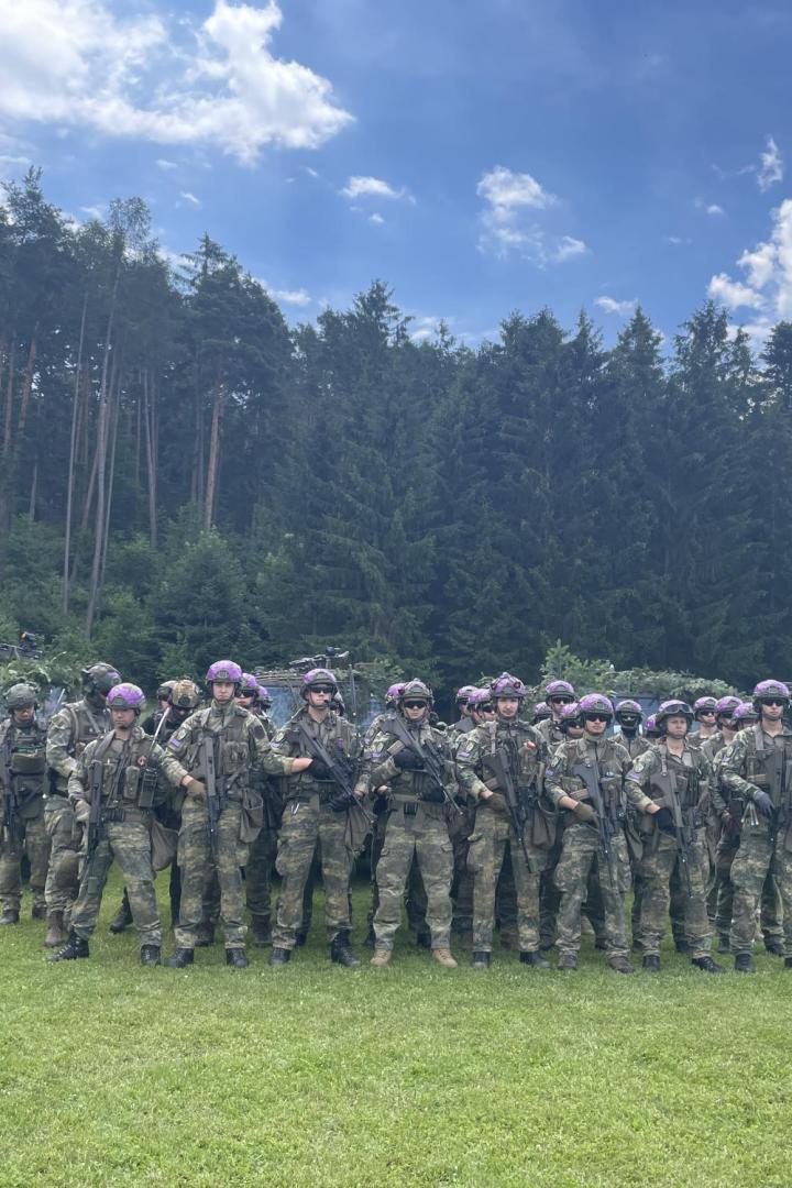 Soldaten mit Waffen posieren vor getarnten Militärfahrzeugen auf einer Wiese.