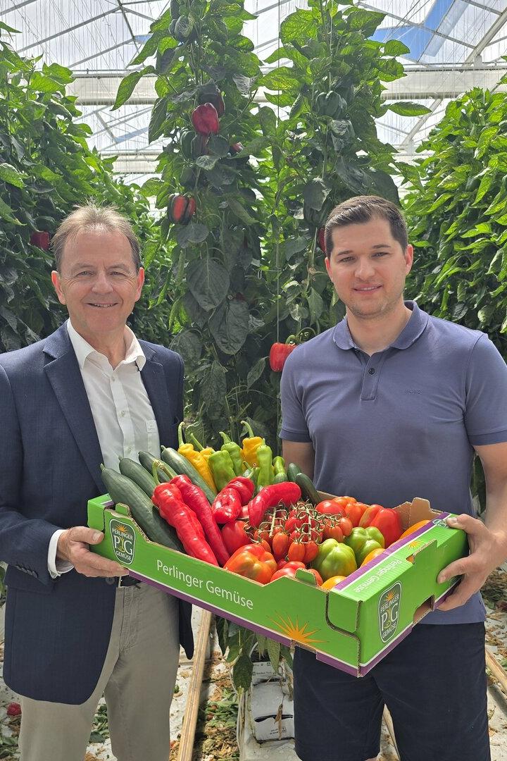 Zwei Männer halten in einem Gewächshaus eine Kiste mit buntem Gemüse vor hohen Paprikapflanzen.