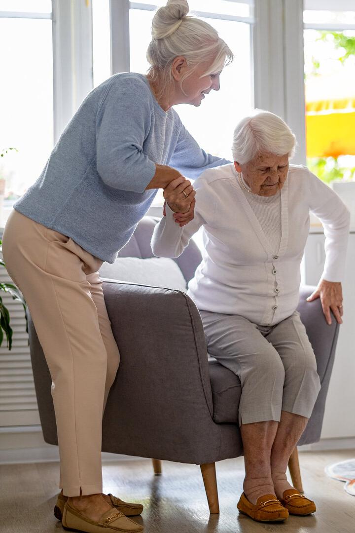 Eine Frau hilft einer älteren Frau beim Aufstehen aus einem Sessel in einem hellen Wohnzimmer.
