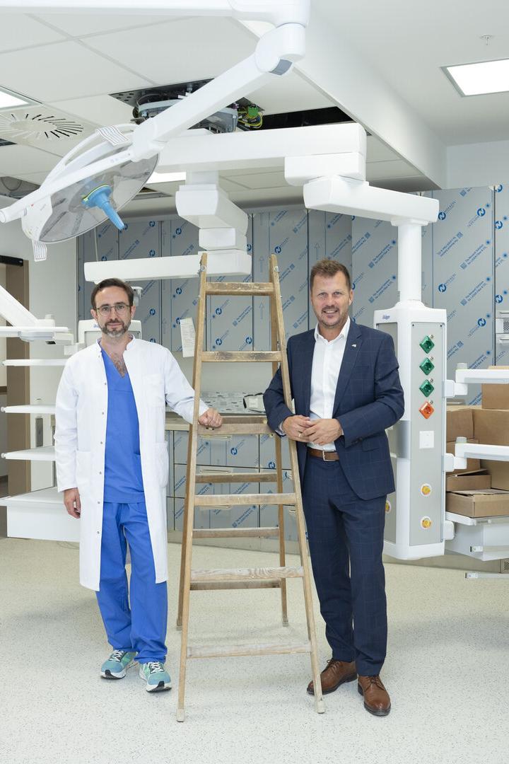 Zwei Männer stehen in einem unvollendeten OP-Raum neben einer Leiter und medizinischer Deckenanlage.