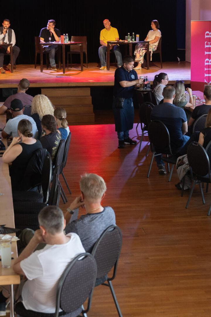 Publikum sitzt bei einer Podiumsdiskussion in einem Saal mit Bühne und roten KURIER-Bannern.