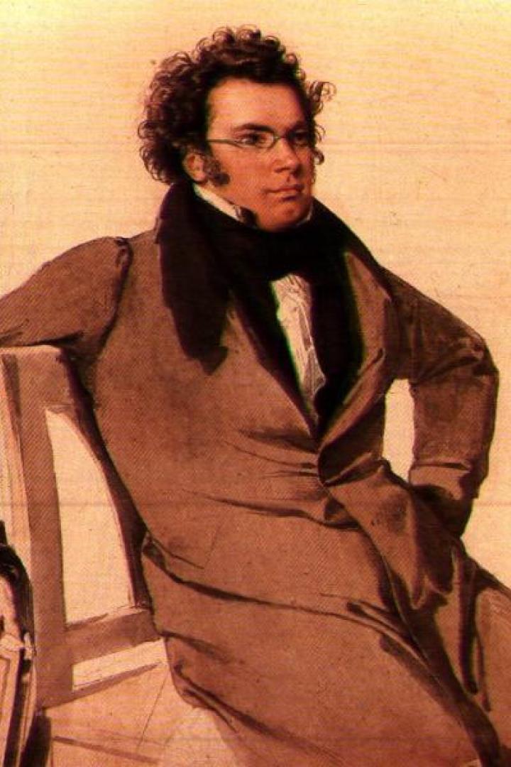 Ein Mann mit Locken, Brille und braunem Mantel sitzt lässig auf einem Stuhl vor hellem Hintergrund.