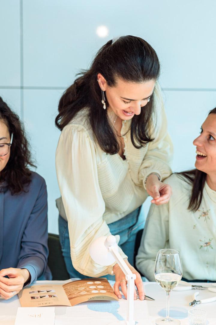 Drei Frauen sitzen an einem Tisch und arbeiten gemeinsam an Schmuckstücken, während eine lächelnd Anweisungen gibt.
