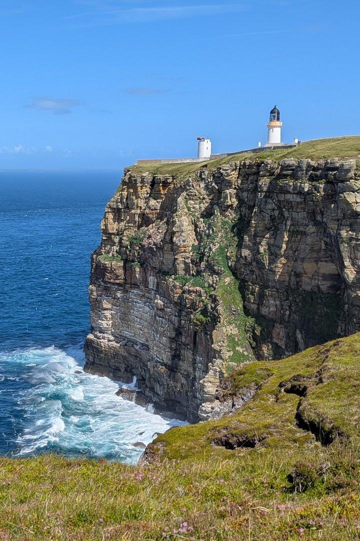 Steilküste am Meer mit Leuchtturm auf der Klippe und Wellenbrandung darunter.