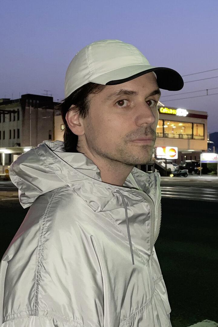 Mann in weißer Kappe und heller Jacke steht an einer nächtlichen Kreuzung mit Ampeln und Bergen im Hintergrund.