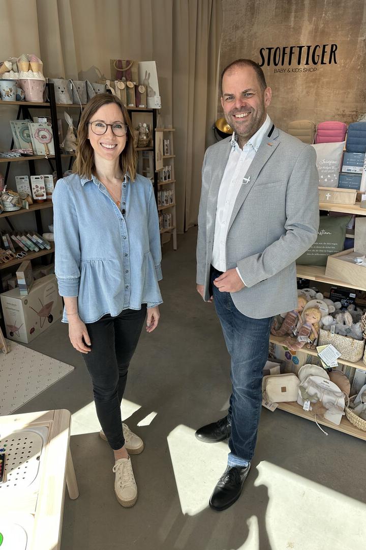 Zwei lächelnde Personen stehen in einem Baby- und Kindershop zwischen Regalen mit Geschenkartikeln und Deko.