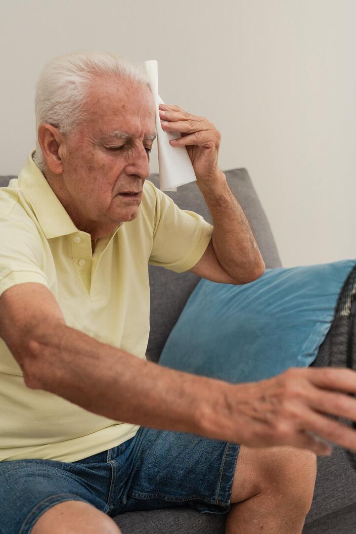 Senior man suffering from summer heat wiping sweat with napkin and holding a electric fan at home