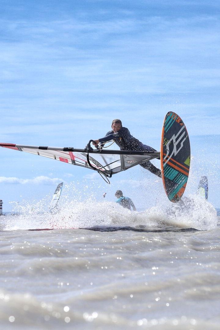 Windsurfer springt über eine Welle im Meer, umgeben von anderen Surfern.