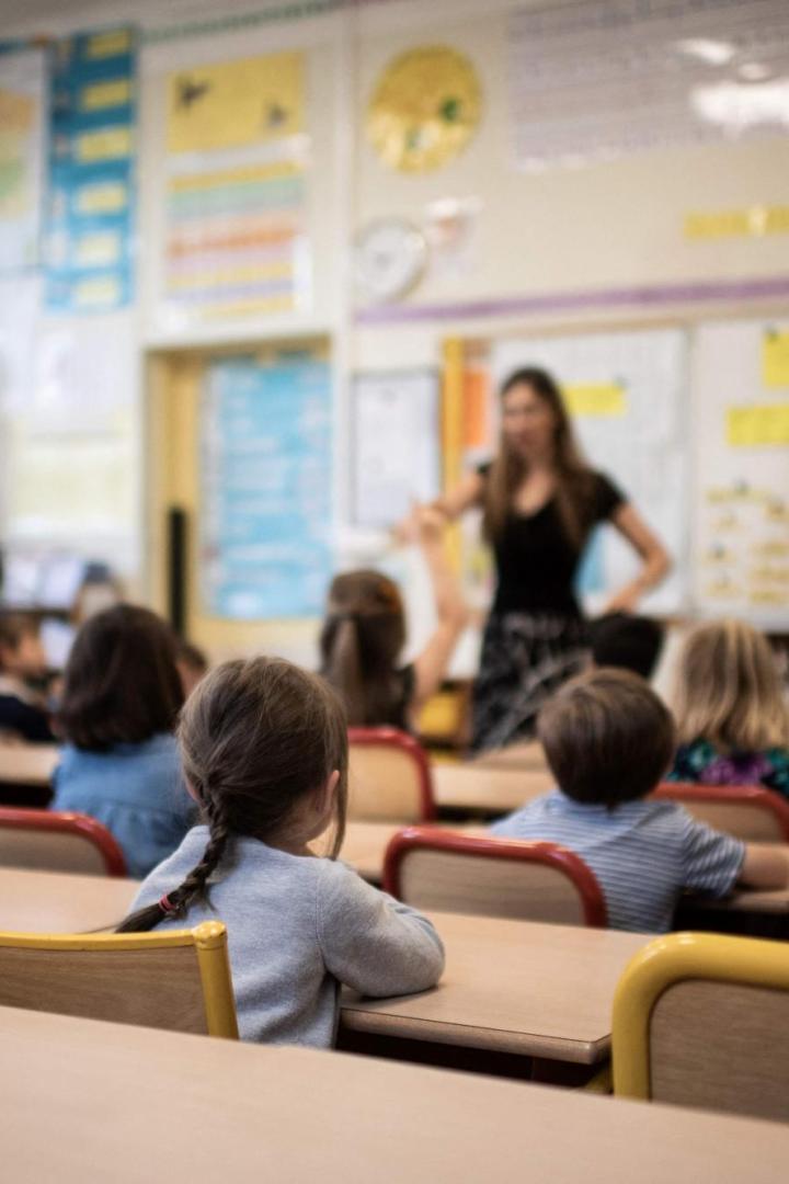Lehrerin erklärt vor einer Grundschulklasse den Stoff, während die Kinder an Tischen sitzen und zuhören.