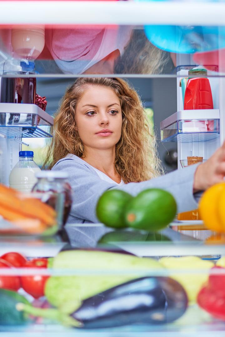 Eine junge Frau mit lockigem Haar greift aus der Perspektive des Kühlschrankinneren nach einem Glas Saft zwischen frischem Obst und Gemüse.