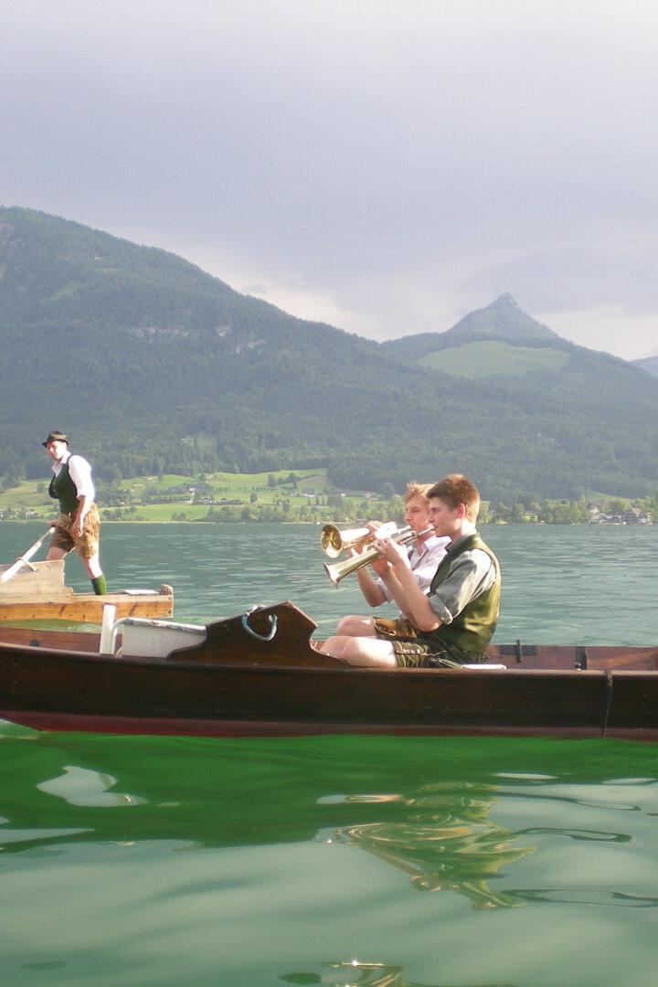 Mehrere Menschen in Tracht fahren und musizieren in Holzbooten auf einem See vor Bergen.