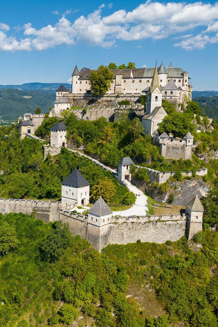 Die Burg Hochosterwitz thront auf einem bewaldeten Felskegel inmitten einer grünen Hügellandschaft in Kärnten.