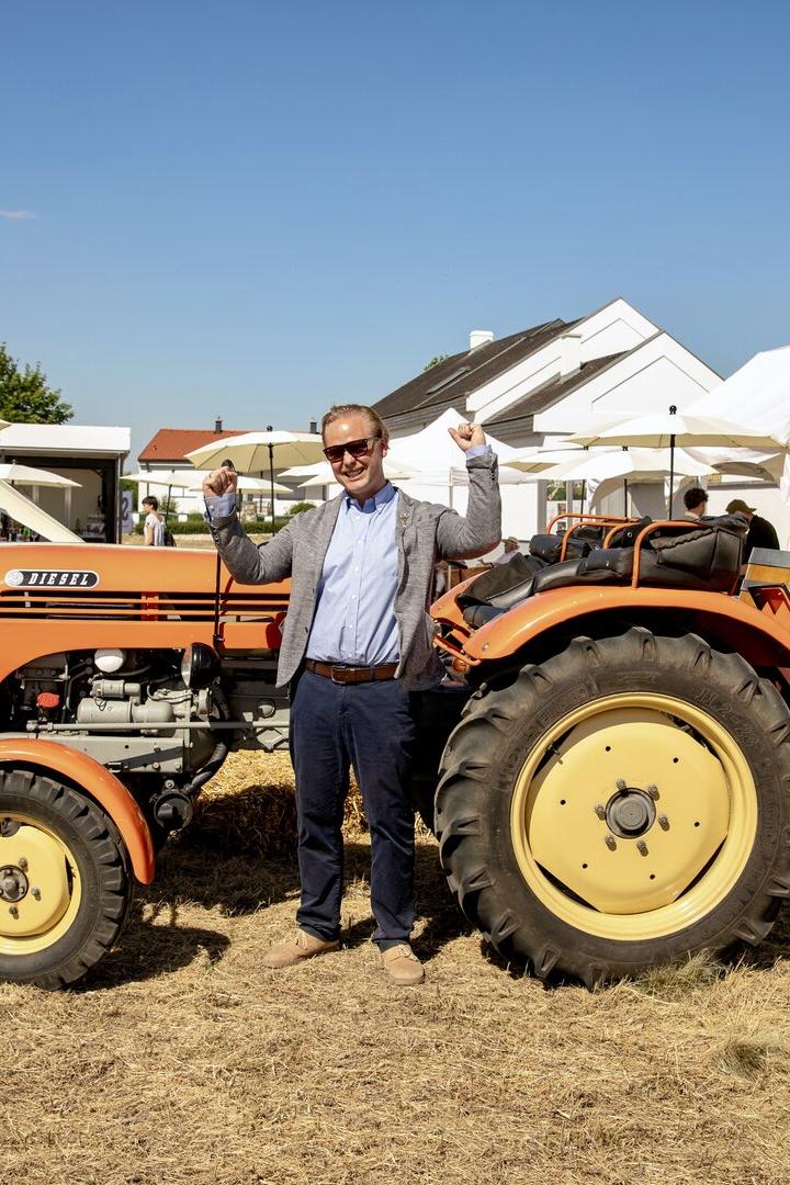Mann posiert lächelnd neben einem orangefarbenen Diesel-Traktor auf einer Feier im Freien.