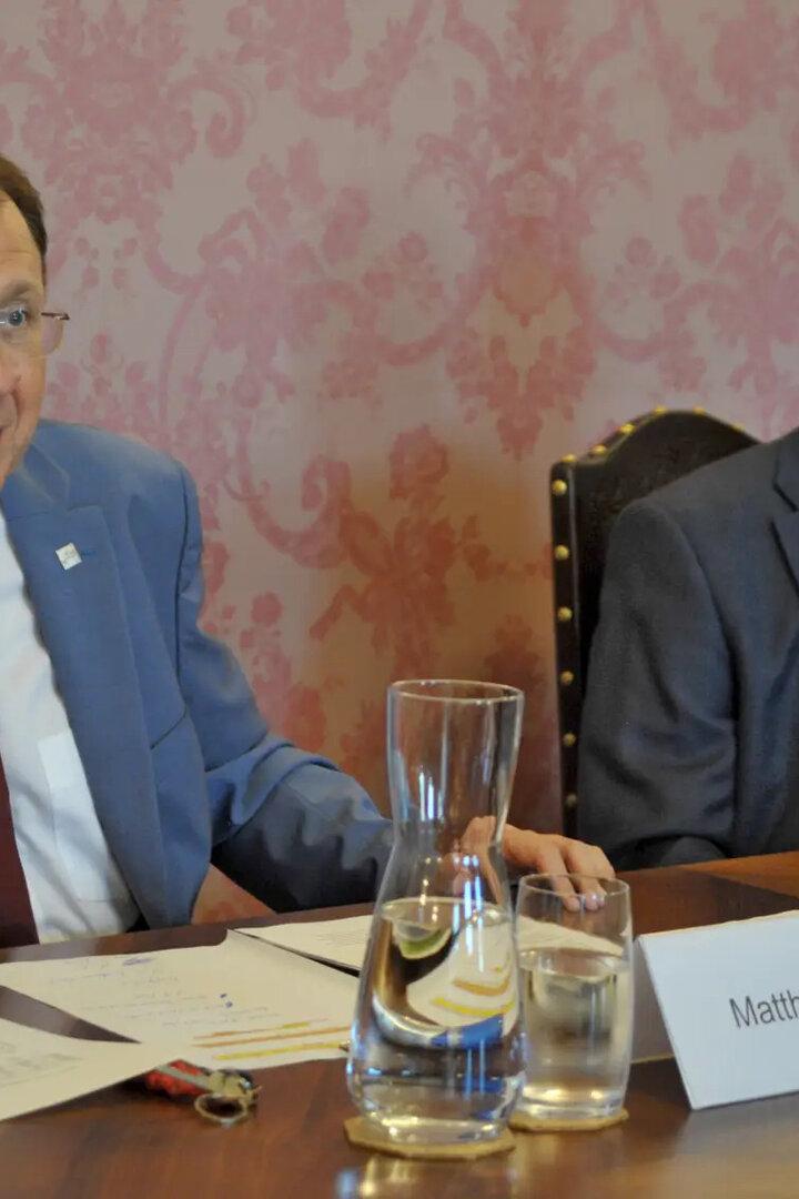 Zwei Männer im Anzug sitzen an einem Konferenztisch mit Wasserkrügen und Namensschild.