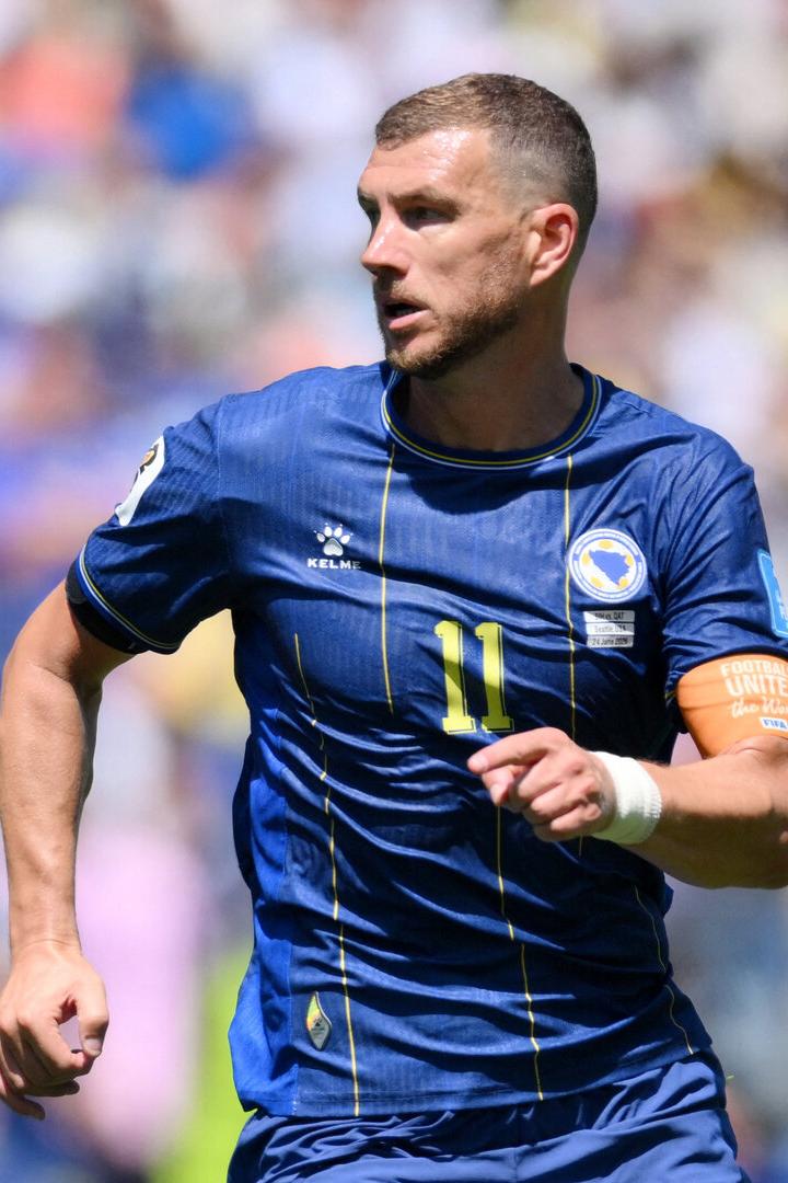 Edin Džeko spielt in einem blauen Trikot der bosnischen Nationalmannschaft mit der Nummer 11 und blickt konzentriert auf einen vorbeifliegenden Fußball.