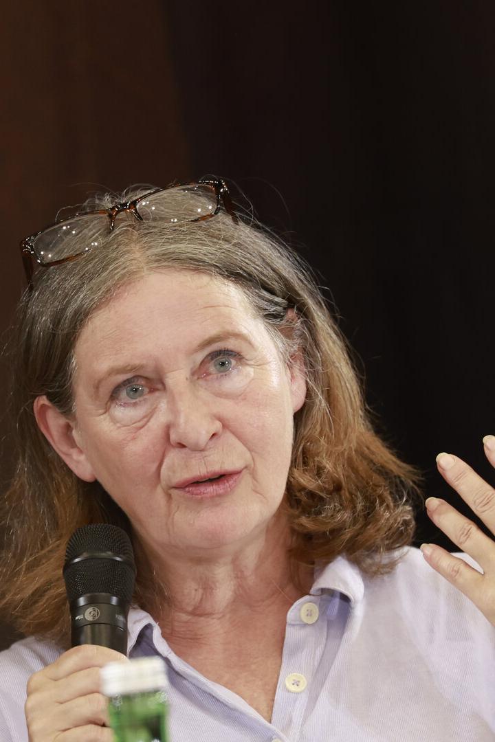 Eine Frau mit Brille auf dem Kopf spricht in ein Mikrofon und gestikuliert mit der Hand vor einem dunklen Hintergrund.