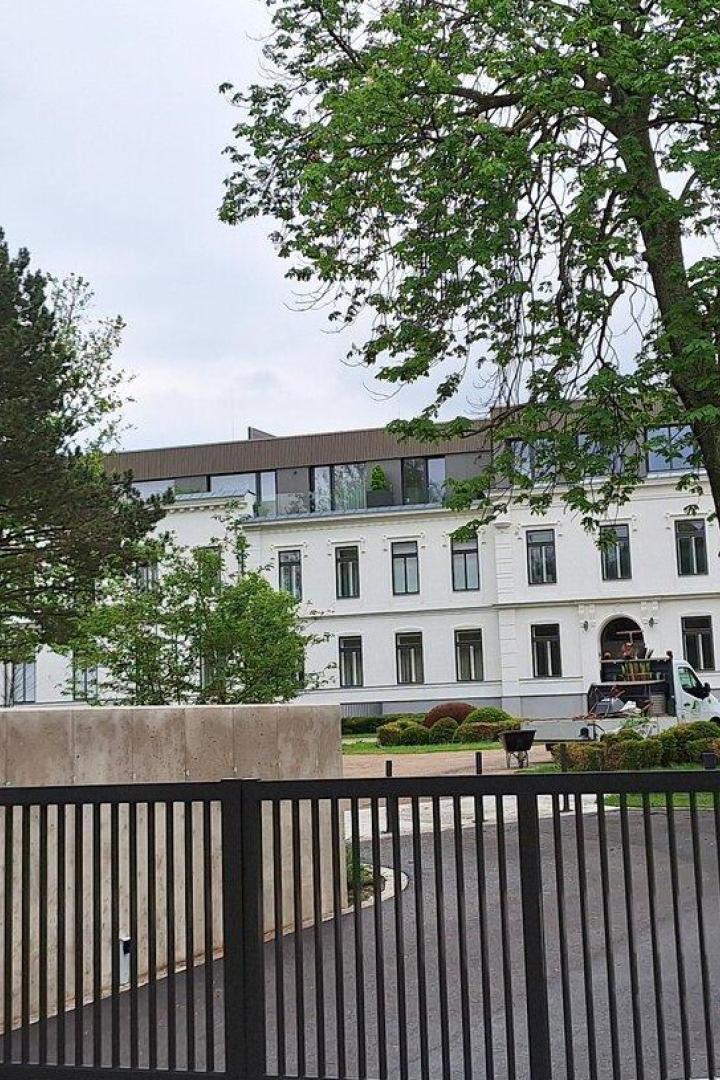 Ein modernes weißes Gebäude hinter einem schwarzen Tor, umgeben von hohen Bäumen und Grün.