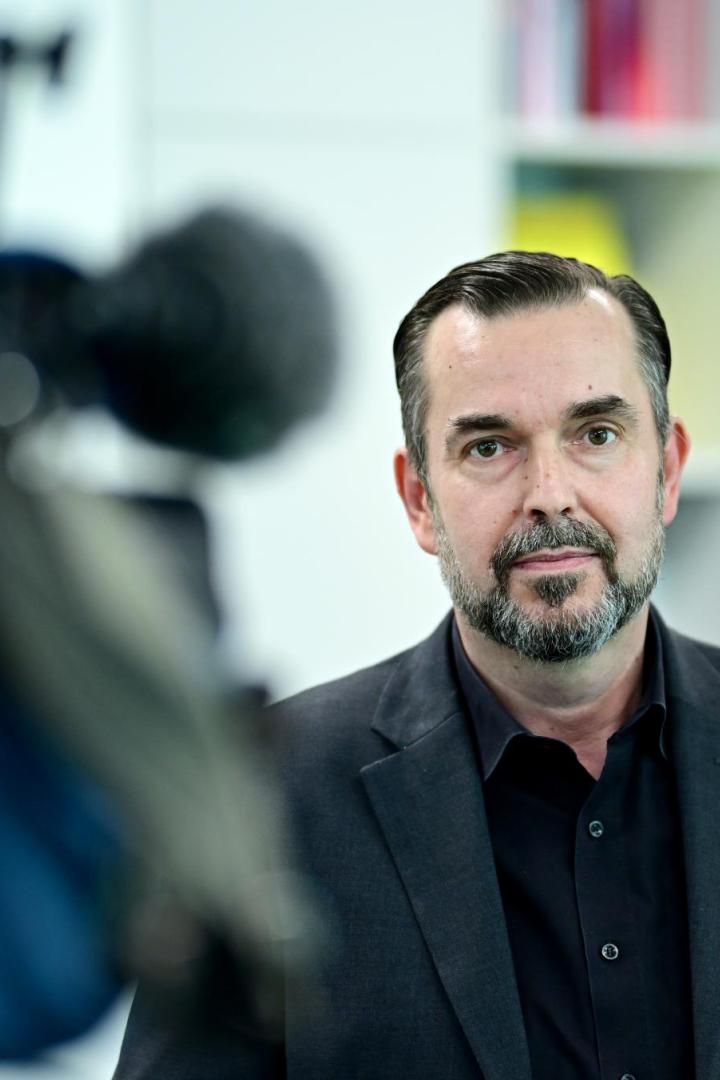 Mann mit Bart im dunklen Anzug blickt in die Kamera, links unscharf eine Filmkamera im Büro.