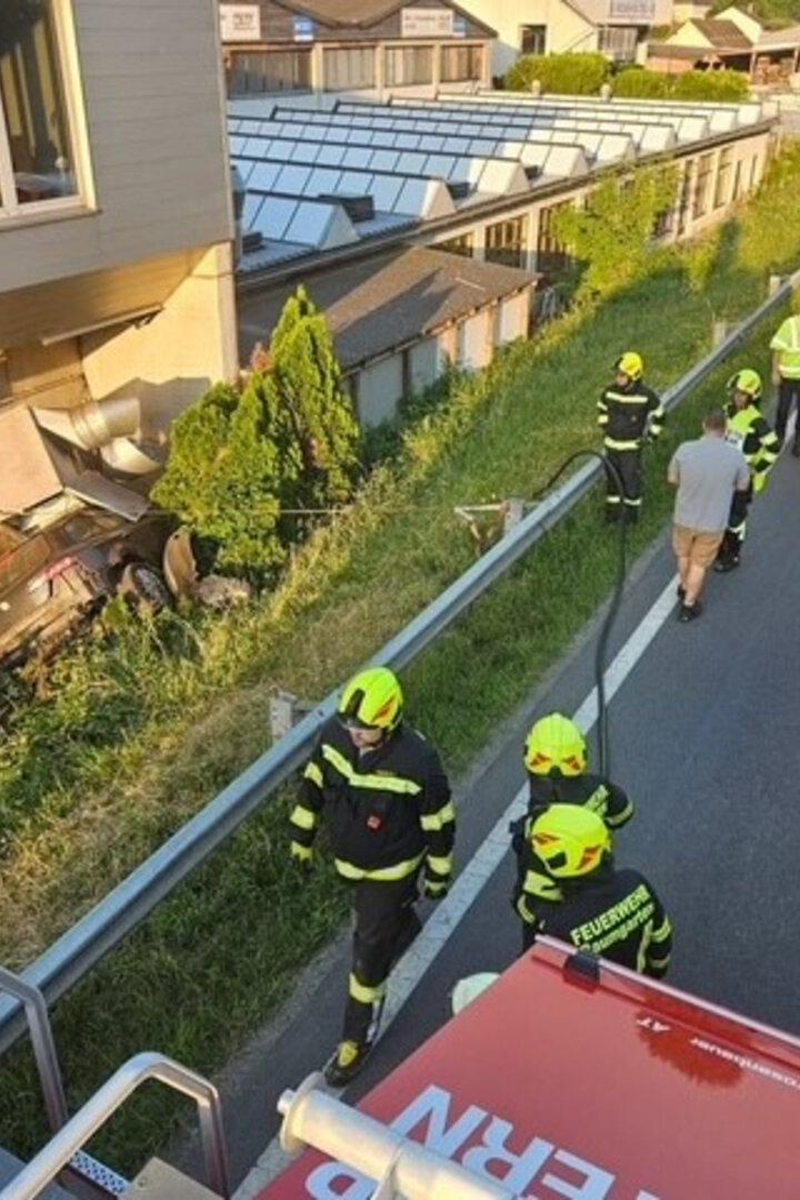 Feuerwehrleute und Rettungskräfte bergen ein Auto an einem Hang neben einem Gebäude; mehrere Einsatzfahrzeuge stehen auf der Straße.