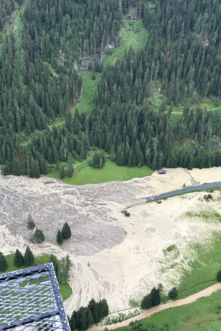 Eine Mure hat sich aus einem bewaldeten Berghang gelöst und eine Schlammlawine über eine Wiese und eine Straße in ein Tal gewälzt.