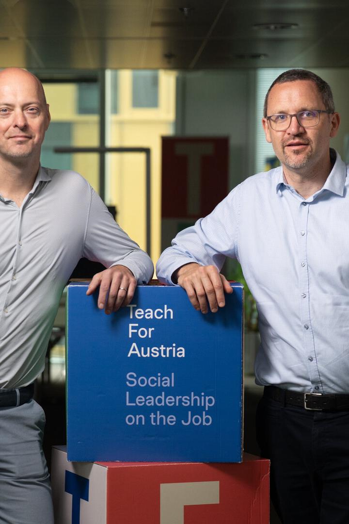Zwei Männer stehen in einem Büroflur neben einem blauen Würfel mit der Aufschrift „Teach For Austria“.