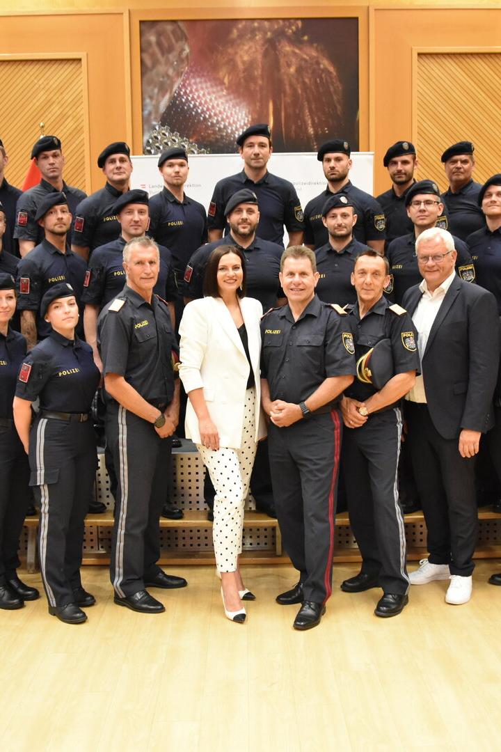 Polizeigruppe in Uniform posiert mit zwei Zivilpersonen in einem Saal vor EU- und Österreich-Flaggen.