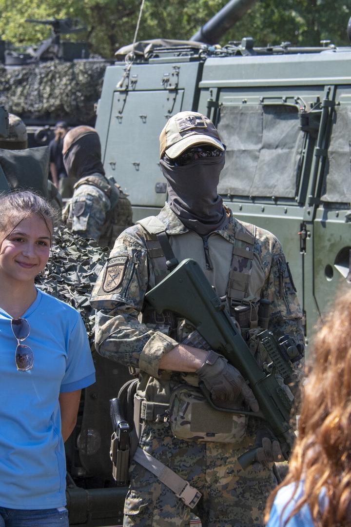 Ein bewaffneter Soldat in Tarnuniform steht vor Militärfahrzeugen, daneben ein lächelndes Mädchen in blauem Shirt.