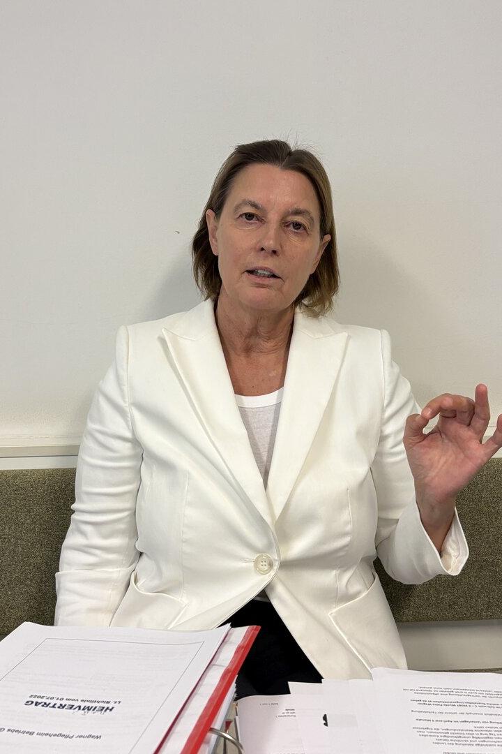 Frau im weißen Blazer sitzt vor Papieren und macht eine Handgeste.