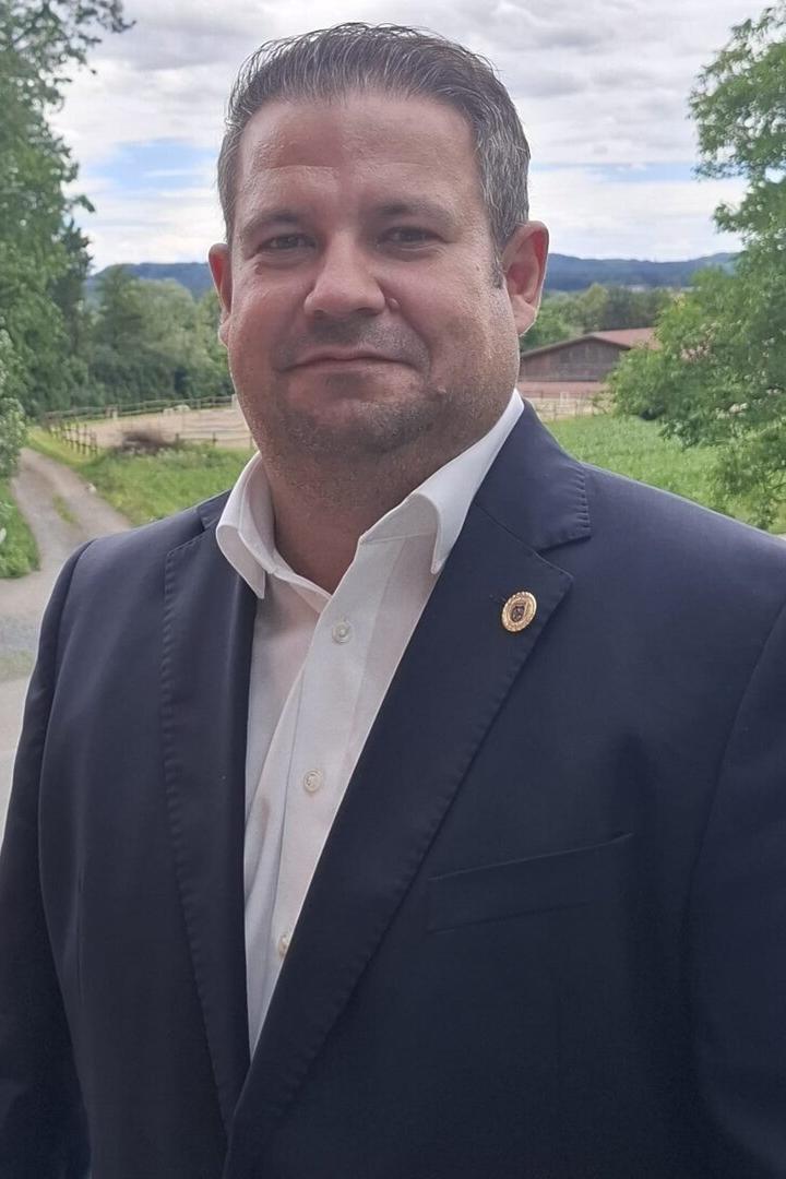 Ein Mann im dunklen Anzug und weißem Hemd steht im Freien vor grüner Landschaft.