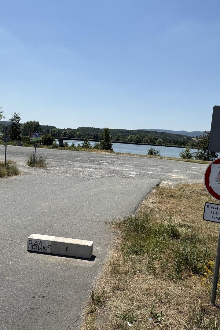 Sperrschilder blockieren eine Straße am Seeufer mit Blick auf Wasser und Hügel.