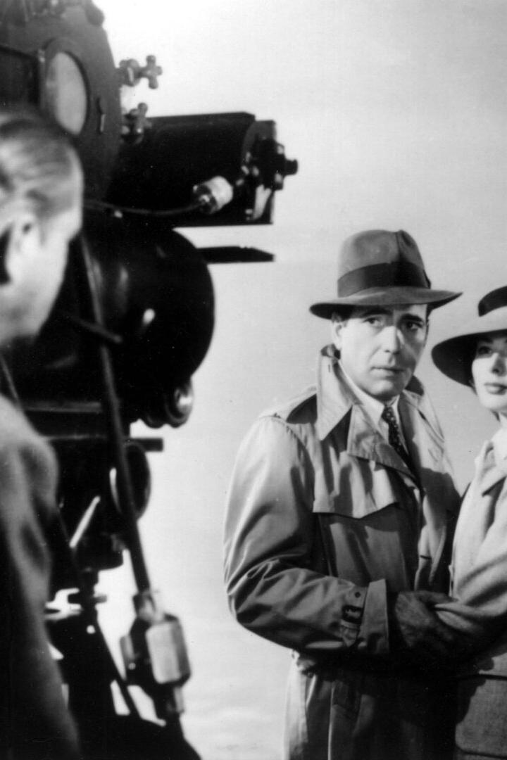 Humphrey Bogart und Ingrid Bergman stehen in Trenchcoats und Hüten vor einer Filmkamera.