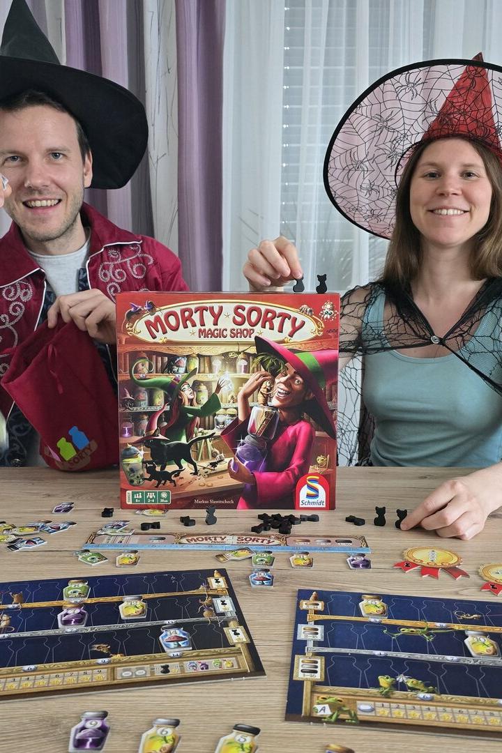 Ein Paar in Hexenkostümen spielt das Brettspiel „Morty Sorty Magic Shop“ an einem Holztisch.
