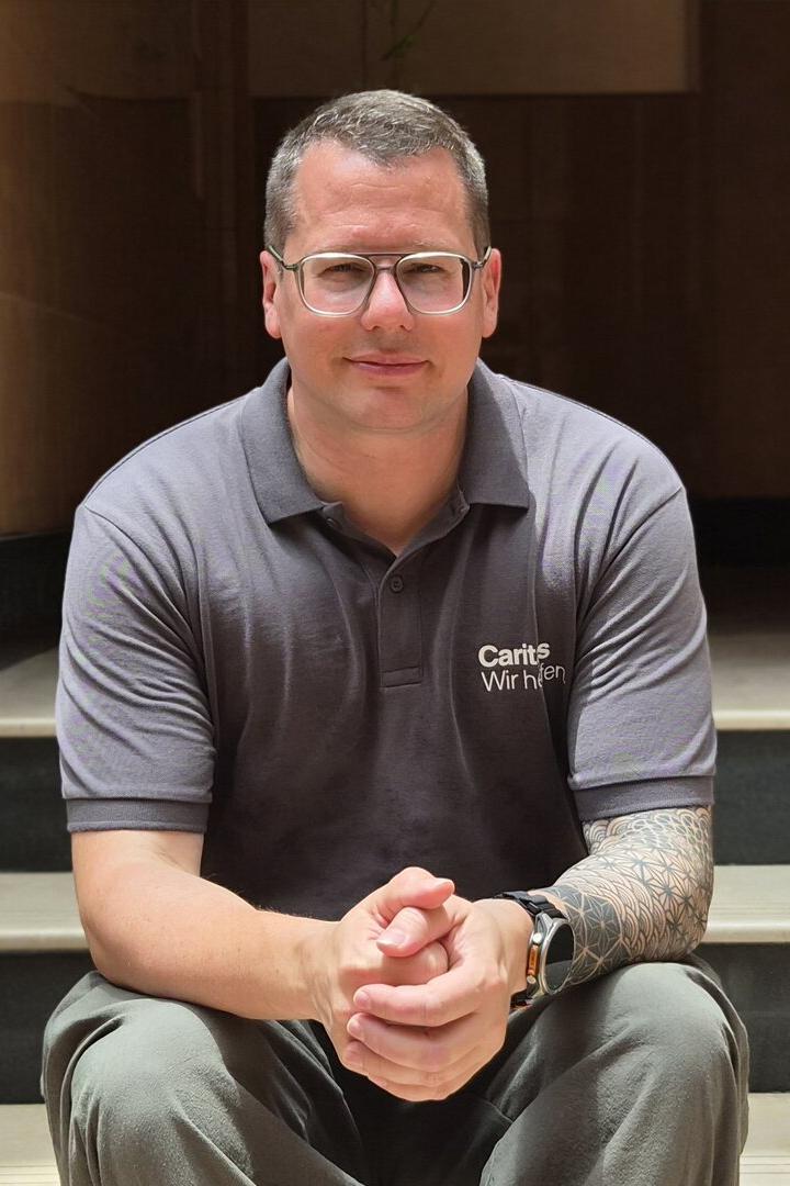 Christoph Riedl mit Brille und Tattoo sitzt auf einer Treppe vor einem dunklen Eingang.