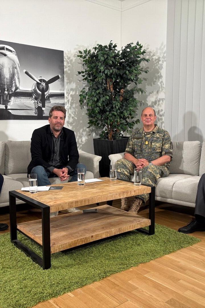 Vier Männer sitzen in einem hellen Sitzbereich auf Sofas um einen Couchtisch, dahinter hängt ein Flugzeugbild.