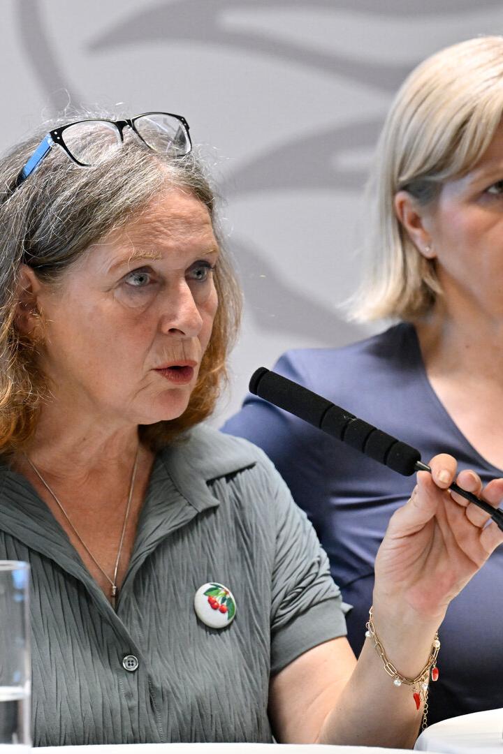 Zwei Frauen bei einer Pressekonferenz; die vordere spricht ins Mikrofon und hält einen Gegenstand hoch.