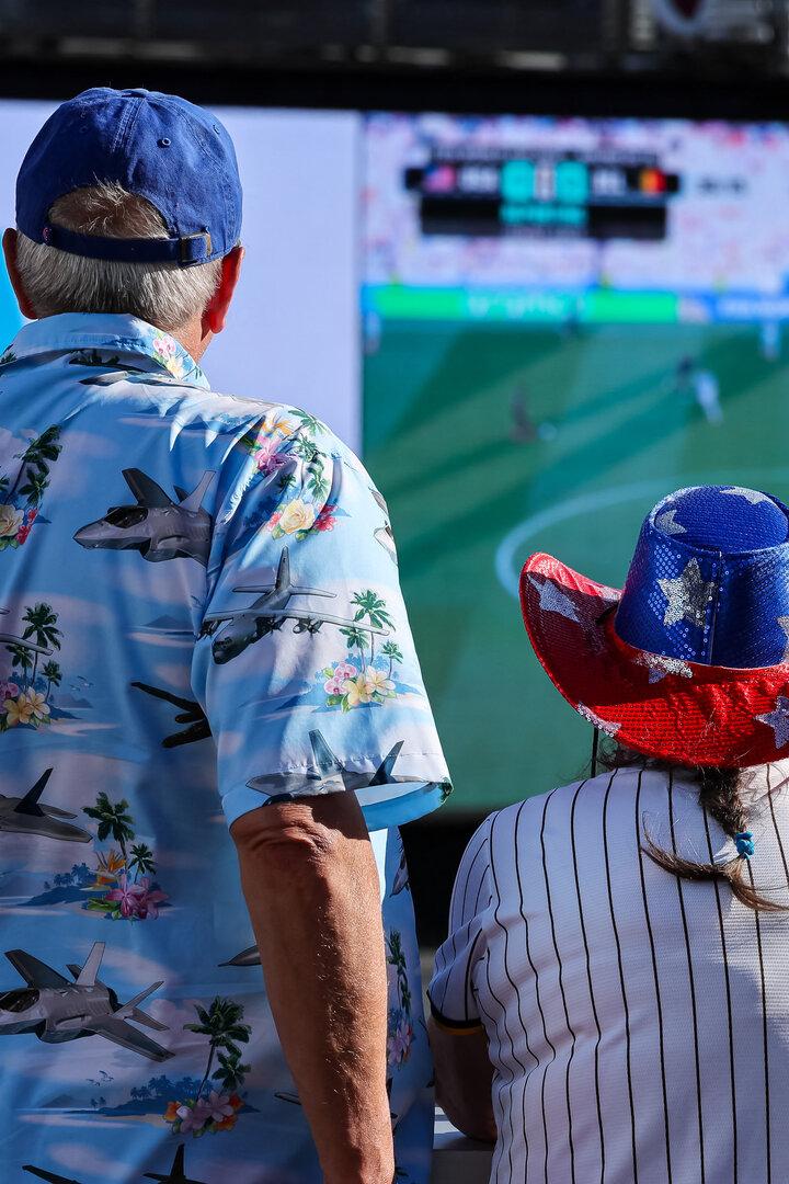 Zwei Personen mit patriotischen Hüten schauen ein Fußballspiel auf einer großen Leinwand.