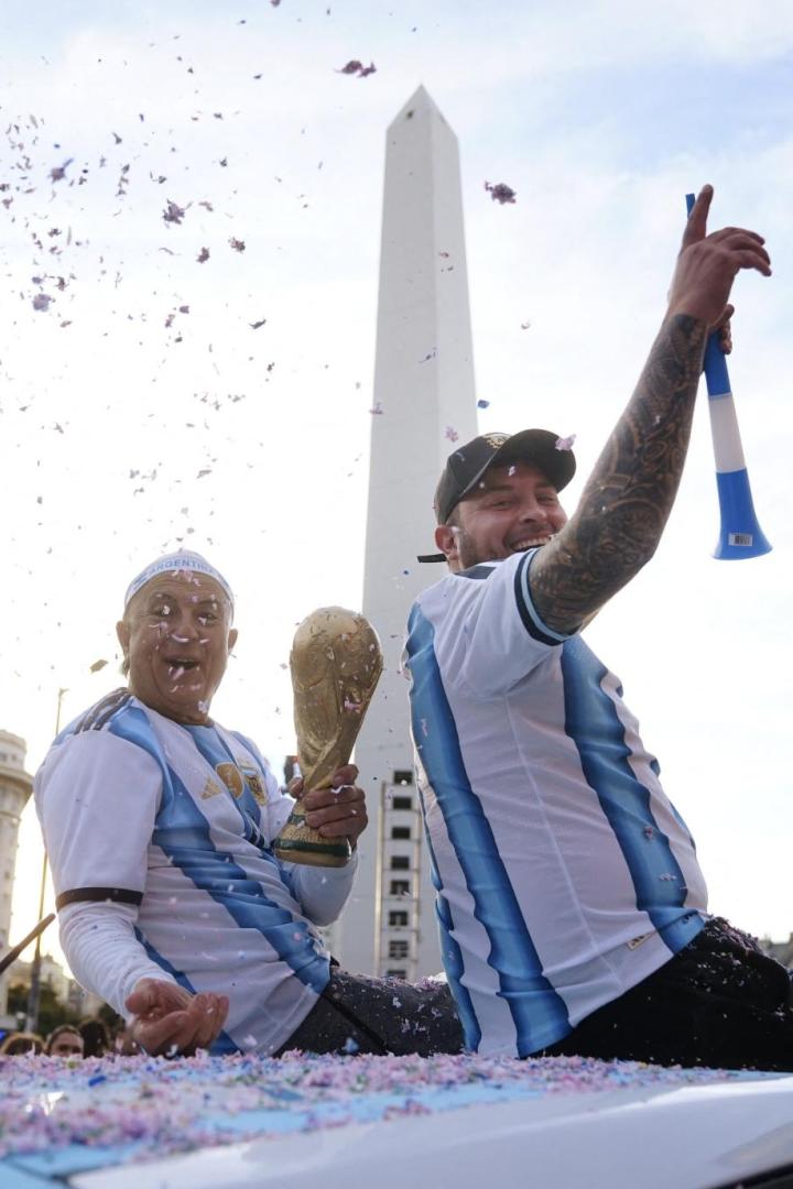 Zwei Fans in Argentinien-Trikots feiern mit dem WM-Pokal vor dem Obelisken in Buenos Aires inmitten von Konfetti.