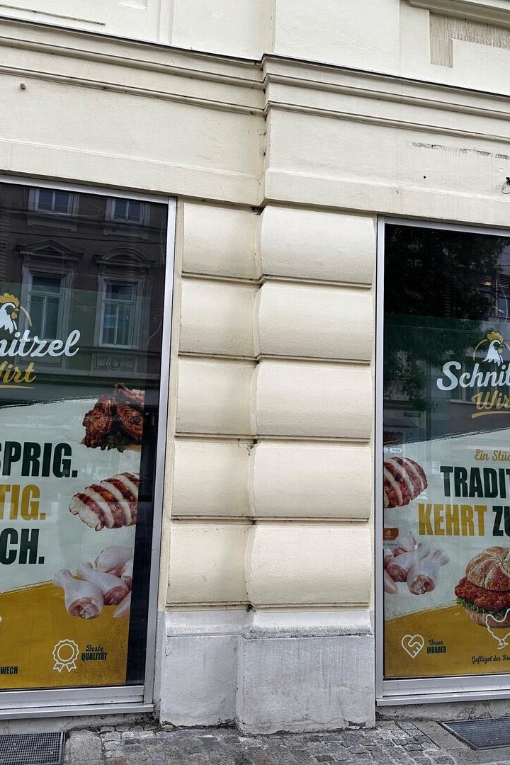 Schaufenster eines Schnitzel-Lokals mit Werbeplakaten für Burger, Hähnchen und Slogans wie „Knusprig. Saftig. Frisch.“.