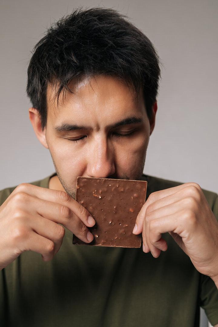 Studio portrait of enjoying young man savoring rich aroma of dark chocolate bar with closed eyes, holding it close to face in anticipation of delightful treat