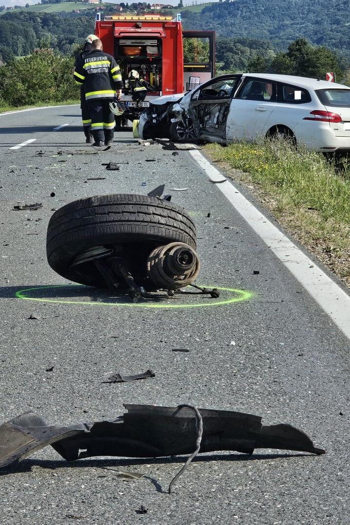 Schwerer Verkehrsunfall mit zerstörtem Auto, Feuerwehr, Trümmern und abgerissenem Rad auf der Straße.
