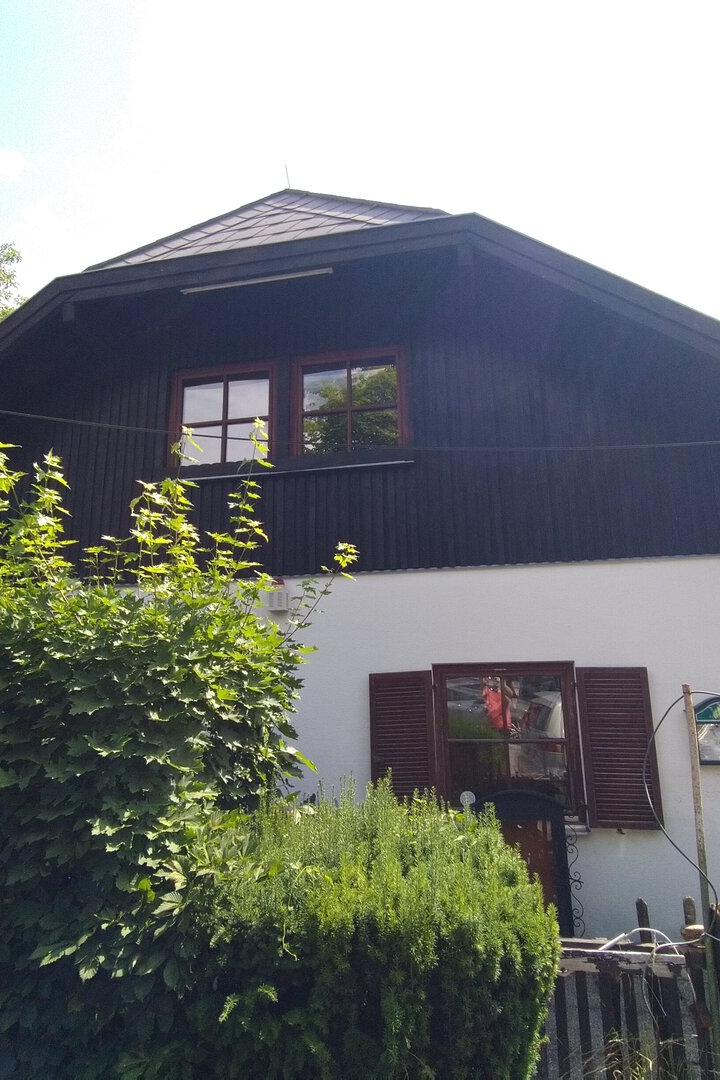Zweistöckiges Haus mit dunklem Dach, weißen Wänden und viel Grün davor, im Vordergrund ein Zaun.