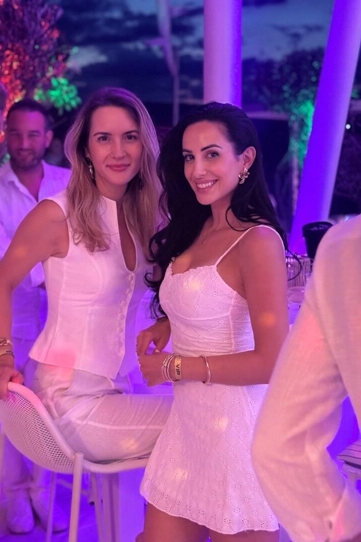 Zwei Frauen in weißen Outfits lächeln bei einer Party im violetten Licht.