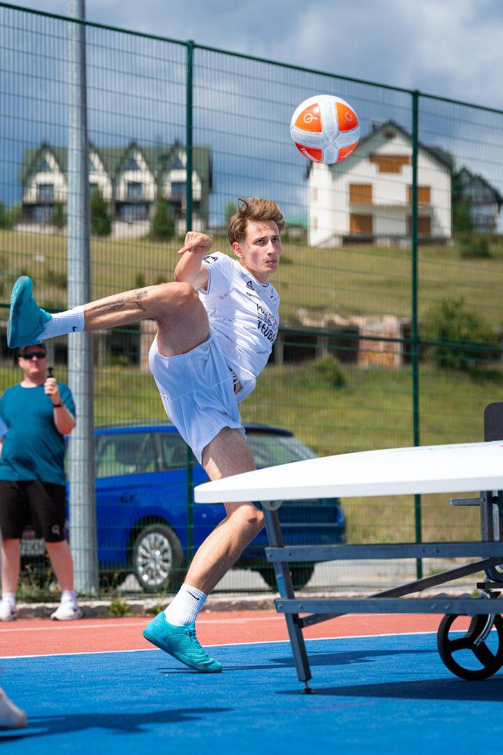 Ein junger Mann in weißer Sportkleidung spielt Teqball auf einem blauen Außenplatz.