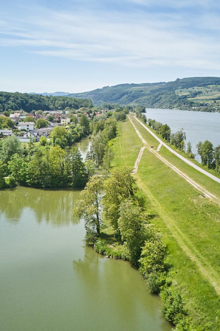 Luftaufnahme eines Flusstals mit Damm, Uferweg, Wohnhäusern und bewaldeten Hügeln.