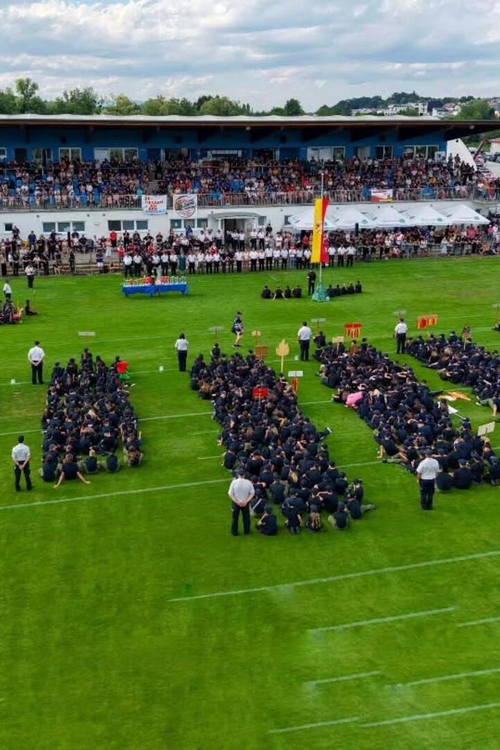 Zahlreiche Feuerwehrleute in Uniform sitzen in Gruppen auf einem Sportplatz vor einer voll besetzten Tribüne bei einer feierlichen Veranstaltung.