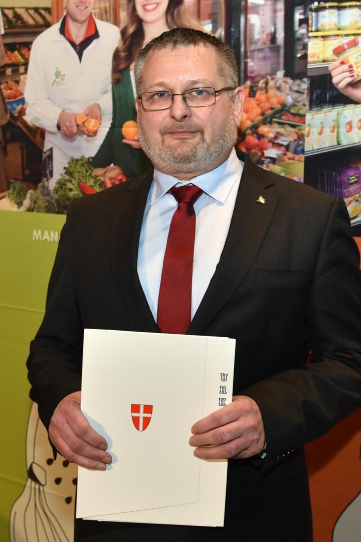 Andreas Kutheil steht vor einer Marktamt-Wandgrafik und hält Unterlagen mit Wiener Wappen in den Händen.